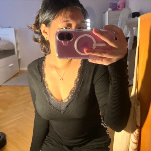 Svart ribbad topp med spets från Amisu - Säljer en svart ribbad långärmad topp från Amisu med v-ringning och fina spetsdetaljer längs halsringningen. Perfekt för att lyfta en enkel outfit med lite extra stil.