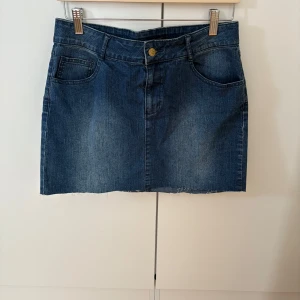 Blå jeanskjol med rå kant - Säljer en klassisk blå jeanskjol med rå, avklippt kant nertill. Kjolen har fem fickor, bälteshällor och stängs med knapp och dragkedja framtill. Perfekt basplagg till garderoben.