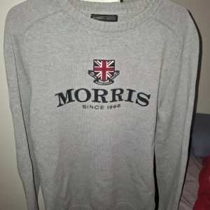 Grå stickad tröja från Morris - Snygg grå stickad tröja från Morris med broderad logga och brittisk flagga på bröstet. Rund halsringning och klassisk passform, perfekt för en stilren look. Tjockt och mjukt material som håller dig varm under kyliga dagar.