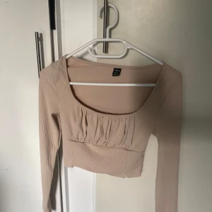 Beige ribbad croppad topp från Gina Tricot - Trendig beige croppad topp från shein  med ribbad struktur och långa ärmar. Toppen har en fyrkantig urringning och rynkade detaljer framtill som ger en snygg touch.