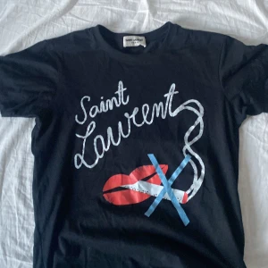 Svart t-shirt från Saint Laurent med tryck - Svart t-shirt från Saint Laurent med coolt tryck framtill. Trycket består av texten 'Saint Laurent', röda läppar och blått/vitt motiv. Klassisk passform och rund halsringning. Perfekt för en trendig look. För frågor om mer bilder eller om passform så är det bara att skriva