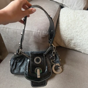 Svart axelväska från Guess med metall-detaljer - Cool svart axelväska från Guess med croco-mönstrad frontficka och stora silvriga metalldetaljer. Väskan har ett stort G-hänge, dragkedja och innerfoder med Guess-logga. Perfekt för dig som gillar statement-accessoarer. Fint skick utöver lädret som är torrt och behöver behandlas! Jag vet ej hur man gör, säljer den därför :(