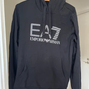 Svart hoodie från Emporio Armani EA7 - Säljer en svart hoodie från Emporio Armani EA7 med stor logga framtill. Tröjan har huva med dragsko och en stor magficka. Perfekt för dig som gillar stilrena och sportiga plagg. Pris kan diskuteras