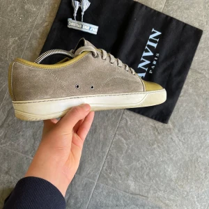 Grå mockasneakers från Lanvin med olivgrön tå - Säljer ett par stilrena sneakers från Lanvin i grå mocka med olivgrön tå i blankt material. Skorna har snörning och vit platt sula. Perfekta för dig som gillar en clean men unik look.