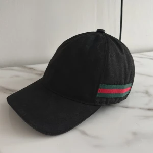 Svart keps från Gucci med röd och grön rand - Säljer en svart keps från Gucci med diskret GG-mönster i tyget och en ikonisk röd och grön rand på sidan. Justerbar rem baktill i svart med gulddetalj. Klassisk böjd skärm och stilren design. (Priset är ej hugget i sten)