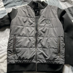 Grå quiltad bomberjacka från Balmain - Säljer en snygg grå bomberjacka från Balmain Paris med quiltad framsida och svarta ärmar. Jackan har dragkedja framtill, två fickor med dragkedja och ribbade muddar. Balmain-logga på bröstet ger en exklusiv känsla.