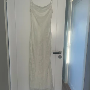 Vit långklänning  - Elegant vit långklänning med tunna axelband och diskret blommönster. Klänningen har en snygg cowlneck-ringning och är figurnära i modellen. Perfekt för mottagning