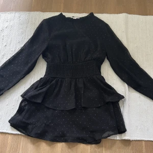 Svart kortklänning med volang från Gina Tricot - Supersöt svart kortklänning från Gina Tricot med små prickar i tyget. Klänningen har lång ärm, volangdetaljer vid kjolen och smockad midja. Perfekt för dig som gillar en feminin och trendig stil.