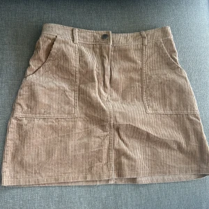 Beige manchesterkjol med fickor - Säljer en beige kortkjol i manchestertyg med två stora fickor framtill och bälteshällor i midjan. Kjolen har en klassisk A-linjeform och stängs med knapp och dragkedja framtill. Perfekt för dig som gillar retrostil!