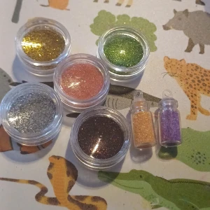 Glittriga lösa pigment i burk och flaska - Flera små burkar och flaskor med lösa pigment i olika färger: guld, grön, rosa, silver, brun, lila och persikofärgad. Perfekt för att skapa skimrande sminklooker eller använda som ögonskugga. Kommer i genomskinliga plastburkar och små flaskor. Dom små är tagna ur 