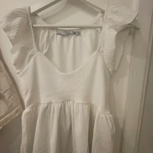 Vit volangtop med u-ringning - Söt vit blis med volangdetaljer på axlarna och u-ringning framtill. Klänningen har en lätt rynkad midja och är tillverkad i ett ribbat material. Perfekt för sommarens alla tillfällen. Köpt på lager 157
