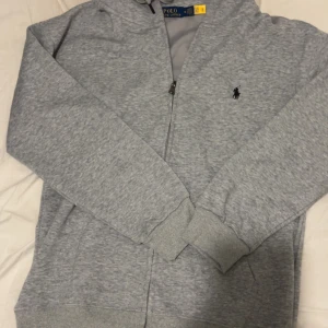 Grå hoodie från Polo Ralph Lauren - Säljer en klassisk grå hoodie från Polo Ralph Lauren med dragkedja och huva. Tröjan har långa ärmar, ribbade muddar och den ikoniska broderade loggan på bröstet. Perfekt för en avslappnad stil.