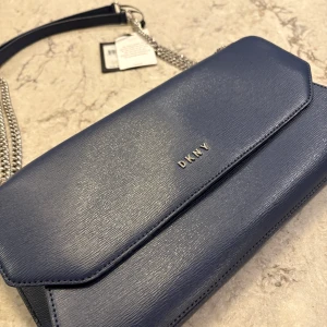 Mörkblå axelväska från DKNY - ✔️Snygg och elegant axelväska från DKNY med justerbar silverfärgad kedjerem och magnetlås.  ✔️Väskan har stilren design med DKNY-logga framtill och är tillverkad i 100% ko skinn ✔️Perfekt storlek för det viktigaste och enkel att bära över axeln. ✔️Väskan är ny med prislapp (den har alltså aldrig använts) ✔️Ny pris på denna var ungefär 2000kr ✔️Har du några frågor/bud/ eller vill se ytterligare bilder så är det bara att höra av dig! 