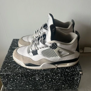 Nike Air Jordan 4 Retro 'Military Black' - Säljer ett par Nike Air Jordan 4 Retro 'Military Black' sneakers. Skorna har en vit ovandel med svarta och grå detaljer, meshpaneler och klassisk snörning. Ikoniska Jumpman-loggor syns på plösen och hälen. Perfekta för dig som gillar streetwear och sneakerskultur.