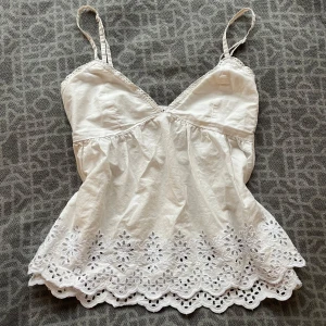Vit broderad linnetopp från BikBok - Intressekoll på en vit linnetopp från BikBok med smala axelband och v-ringning. Toppen har broderade blommor och hålmönster nertill samt vågig kant. .