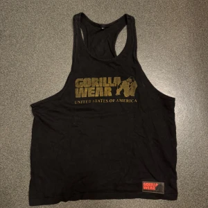 Svart linne Gorilla Wear - Ett svart linne från Gorilla Wear med vanlig passform. Guldfärgad text fram och diskret logga bak. Perfekt linne för gymmet.