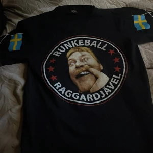 Svart t-shirt med tryck och svenska flaggor - Svart t-shirt med stort tryck på bröstet med texten 'RUNKEBALL RAGGARDJÄVEL' och ett ansikte i mitten. Svenska flaggor på båda ärmarna. Rund halsringning och normal passform.