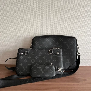 Axelväska med monogrammönster - Säljer en snygg axelväska i svart och grått med klassiskt monogrammönster. Väskan har flera delar, justerbar axelrem och silverfärgade metalldetaljer. Perfekt för dig som vill ha en stilren och lyxig accessoar.