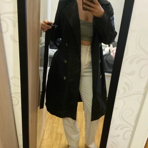 Svart trenchcoat med bälte - Säljer en klassisk svart trenchcoat med knappar och tillhörande bälte i midjan. Jackan har lång ärm och en rak, längre passform som går ner mot knäna. Perfekt för dig som vill ha en stilren och tidlös jacka.
