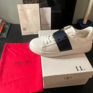 Valentino Garavani Open - Säljer ett par vita Valentino Garavani Open sneakers med bred blå läderdetalj över mitten. Skorna har snörning, rund tå och platt sula. Klassisk och stilren design med diskret Valentino-logga på plösen. Aldrig använda. Helst byten mot andra skor i 45. Skriv gärna om du har nån fråga⭐️