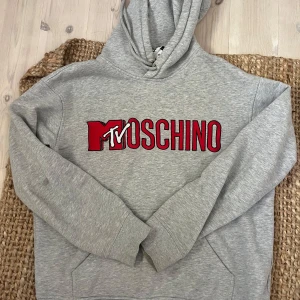 Grå hoodie från Moschino x MTV - Säljer en grå hoodie från Moschino i samarbete med MTV. Tröjan har en stor röd och vit logga framtill, huva med dragsko och magficka. Perfekt för dig som gillar streetwear och vill sticka ut med en ikonisk logga.