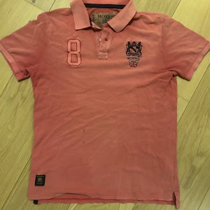 Rosa pikétröja från Morris - Säljer en rosa pikétröja från Morris med klassisk krage och knappslå. Tröjan har broderad logga och brittisk flagga på bröstet samt en stor siffra 8. Perfekt för en stilren och sportig look.