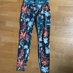  Träningstights från Stronger - Supersnygga träningstights från Stronger med färgglatt blommönster i rött, vitt och grönt. Hög midja med logotyp och två gröna ränder på ena benet. Perfekta för gymmet eller löprundan.