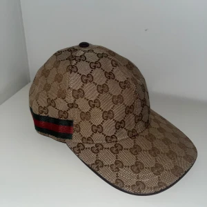 Beige keps från Gucci med GG-mönster - Säljer en beige keps från Gucci med klassiskt GG-monogram över hela kepsen. Kepsen har en brun läderrem baktill för justering och en röd-grön rand på sidan. Tillverkad i bomullsmaterial med detaljer i läder.
