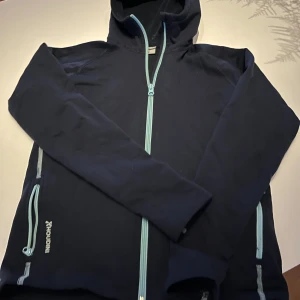 Mörkblå hoodiejacka från Houdini st 150 - Mörkblå hoodiejacka från Houdini med ljusblå dragkedjor och huva. Jackan har långa ärmar och två fickor med dragkedja. Perfekt för lager-på-lager och chill dagar.