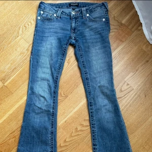 Blå bootcut jeans från True Religion - Säljer ett par klassiska blå bootcut jeans från True Religion med snygga kontrastsömmar och ikoniska bakfickor. Jeansen har normal passform och stängs med knapp och dragkedja. Perfekta för dig som gillar en tidlös jeanslook.