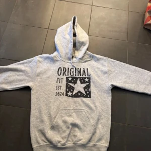Scndi hoodie - Säljer en grå hoodie med svart tryck framtill där det står 'ORIGINAL FIT EST. 2024' och en stor stjärna med mindre stjärnor runtom. Tröjan har huva med dragsko och en stor magficka. Perfekt för en avslappnad stil.
