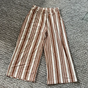 Brun- och vitrandiga byxor från Gina Tricot - Vida byxor från Gina Tricot i brun- och vitrandigt mönster. Byxorna har resår i midjan och är tillverkade i en mjuk viskosblandning. Perfekta för en avslappnad och trendig stil.