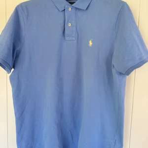 Ljusblå pikétröja från Polo Ralph Lauren - Säljer en klassisk ljusblå pikétröja från Polo Ralph Lauren med gul broderad logga på bröstet. Tröjan har krage, två knappar och korta ärmar. Perfekt för en stilren och avslappnad look.
