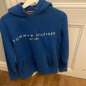 Blå hoodie från Tommy Hilfiger - Säljer en blå hoodie från Tommy Hilfiger med vit broderad logga och klassisk flagga framtill. Tröjan har huva och en stor magficka. Perfekt för dig som gillar stilrena och bekväma plagg.