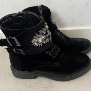 Svarta boots med strassdetaljer och spännen - Säljer ett par svarta boots med glittriga strassdetaljer och dubbla spännen på sidan. Skorna har snörning framtill och dragkedja på insidan. Perfekta för dig som vill sticka ut med lite extra bling!