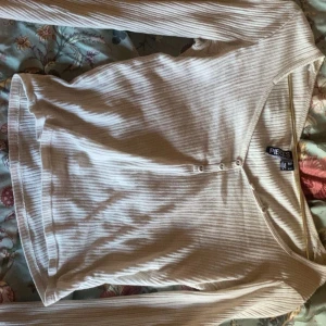 Beige ribbad omlottopp från Pieces - Säljer en beige ribbad omlottopp från Pieces med lång ärm och knappar framtill. Toppen har en snygg v-ringning och är i mjukt material som sitter skönt på kroppen.