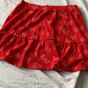 Röd kjol med volang från H&M Divided - Säljer en röd kjol från H&M Divided med vita blommönster och volangdetalj. Kjolen är kort och har en luftig känsla, perfekt för varma dagar. Resår i midjan för bekväm passform.