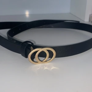 Svart bälte med guldfärgat spänne - Säljer ett stilrent svart bälte med ett snyggt guldfärgat dubbelring-spänne. Perfekt accessoar för att lyfta din outfit. Bältet är smalt och gjort i ett slätt material.