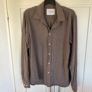 Brun skjorta från Les Deux - Säljer en brun långärmad skjorta från Les Deux med klassisk krage och vita knappar. Skjortan har diskret broderad logga på bröstet och är tillverkad i ett mjukt material som känns bekvämt mot huden. Storlek M (pris kan diskuteras)