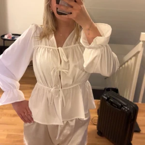 Vit blus med knyt och volang - Säljer en söt vit blus med knyt framtill och volangdetaljer vid ärmslut och midja. Blusen har långa ärmar och en lös passform, perfekt för en trendig och avslappnad look.