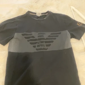 Svart t-shirt från Emporio Armani EA7 - Säljer en svart t-shirt från Emporio Armani EA7 med stort logotryck på bröstet och guldfärgad EA7-logga på ärmen. Klassisk passform och rund halsringning. 