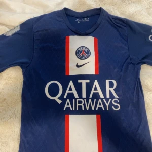 PSG hemmatröja 2022/23 med nummer 10 - Säljer en blå Paris Saint-Germain fotbollströja från Nike med Qatar Airways som sponsor och nummer 10 på ryggen. Tröjan har korta ärmar, klassisk V-ringning och vita samt röda detaljer på framsidan. Perfekt för fans av PSG!