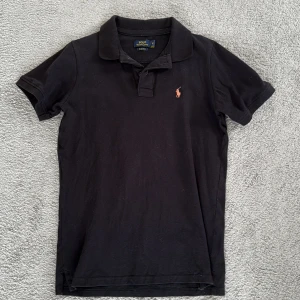 Svart pikétröja från Polo Ralph Lauren äkta storlek M  - Klassisk svart pikétröja från Polo Ralph Lauren med korta ärmar och krage. Tröjan har den ikoniska broderade loggan i orange på bröstet och är tillverkad i bomull. Perfekt för en stilren och avslappnad look.