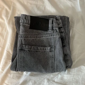 Grå jeans från RYVLS - Säljer ett par grå jeans från RYVLS med klassisk femficksdesign och svarta detaljer. Jeansen har raka ben och normal passform, perfekta för en stilren vardagslook. Snyggt svart läderpatch bak i midjan.