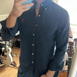 Mörkblå skjorta från Ralph Lauren - Säljer en mörkblå skjorta från Ralph Lauren med klassisk krage och knappar framtill. Slim fit-modell med lång ärm och ikonisk röd broderad logga på bröstet. Perfekt för dig som gillar stilrena och tidlösa plagg.