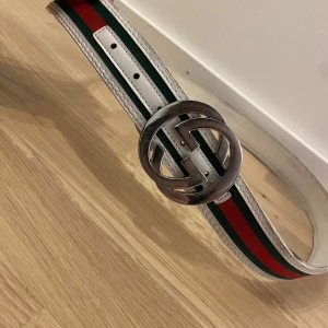 Gucci bälte - Snyggt bälte från gucci. Bältet har vit bas med gröna och röda ränder längs hela bandet. Tillverkat i skinn med tydliga sömmar och bred design. Perfekt accessoar för att lyfta din outfit. Bältet är 115 cm. Väldigt snyggt bälte som ger en lyxig känsla. Vintage gucci bälte. 