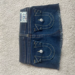 Mörkblå jeanskjol från True Religion - Säljer en mörkblå jeanskjol från True Religion med rå kant nertill och klassiska kontrastsömmar. Kjolen har två stora bakfickor med lock och knapp samt detaljerade sömmar. Perfekt för dig som gillar street och denim.