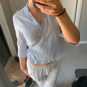 Ljusblå omlottblus med volangdetaljer - Säljer en ljusblå omlottblus med tunna vita ränder och volang längs omlottkanten. Blusen har trekvartsärm och knyt i sidan, vilket ger en feminin och avslappnad look. Perfekt för vår och sommar!