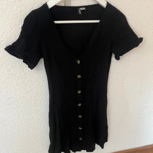 Svart kortklänning med knappar från H&M - Svart kortklänning från H&M med v-ringning och knappar framtill. Klänningen har korta ärmar och en figurnära passform. Perfekt för dig som gillar enkel och stilren design.💝💝 Säljer då den är för liten för mig! 