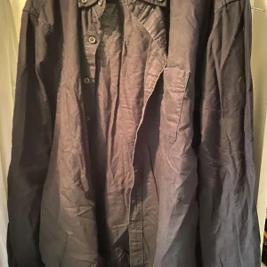 Svart skjorta i bomull - Säljer en svart långärmad skjorta med klassisk krage och knäppning framtill. Skjortan har en bröstficka och är tillverkad i ett mjukt bomullstyg. Perfekt för en stilren och enkel look.